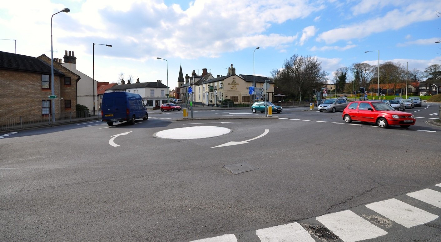 a UK road junction with two small flat painted mini-roundabout circles connected by a short two-way link road, with cars navigating both.