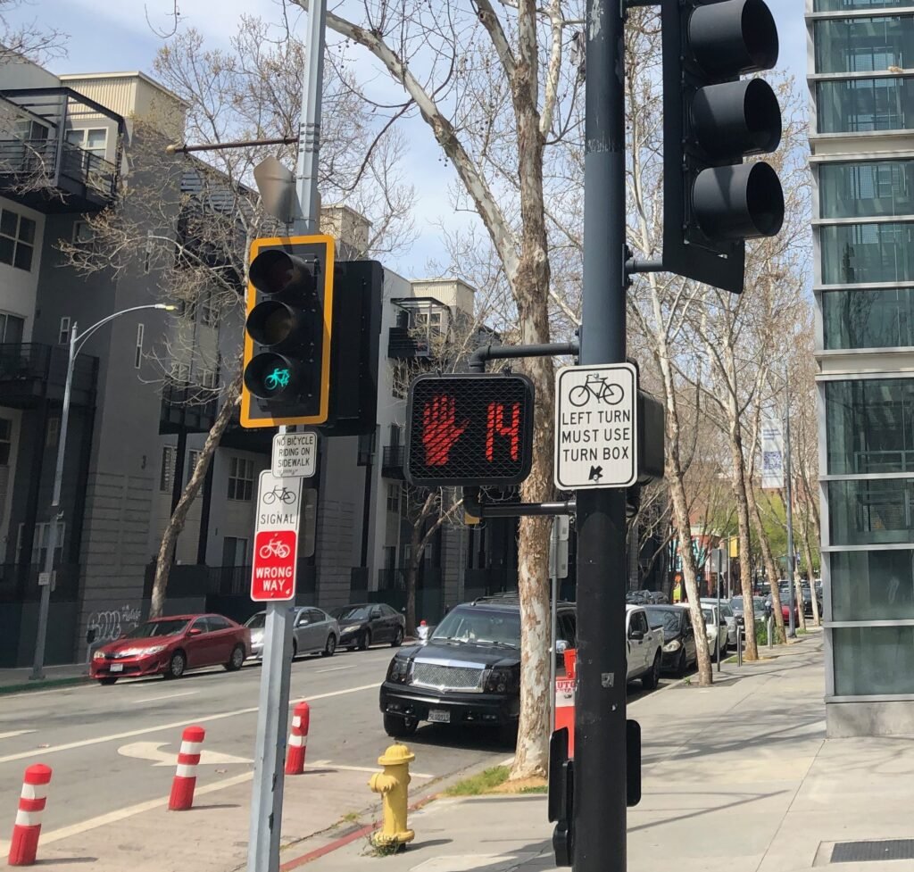 Green bicycle signal beside a pedestrian countdown display and bicycle left-turn instruction sign in San Jose.