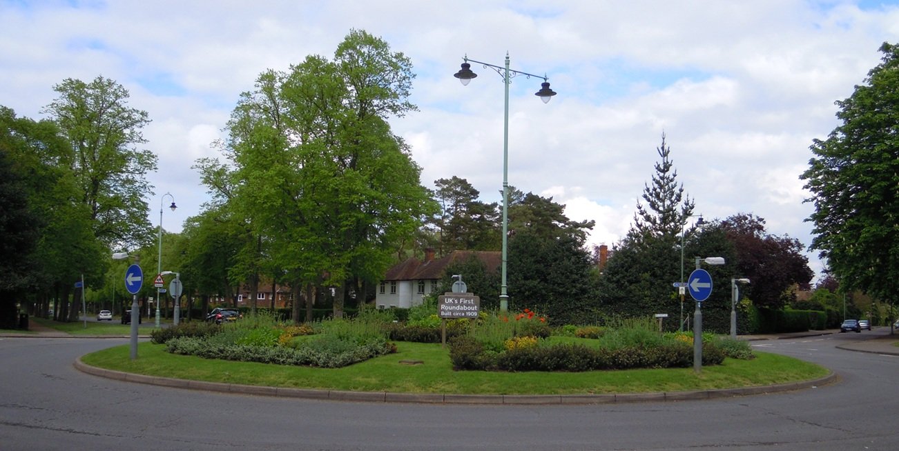 photograph of a small early 20th-century circular road junction with a central island, a few period cars, and low buildings in the background.