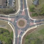 Aerial view of a four-leg, multi-lane modern roundabout. The design features a landscaped central island surrounded by a reddish mountable truck apron, raised splitter islands on each approach, high-visibility pedestrian crosswalks set back from the intersection, and pavement markings indicating yield lines and circulating lane directions.