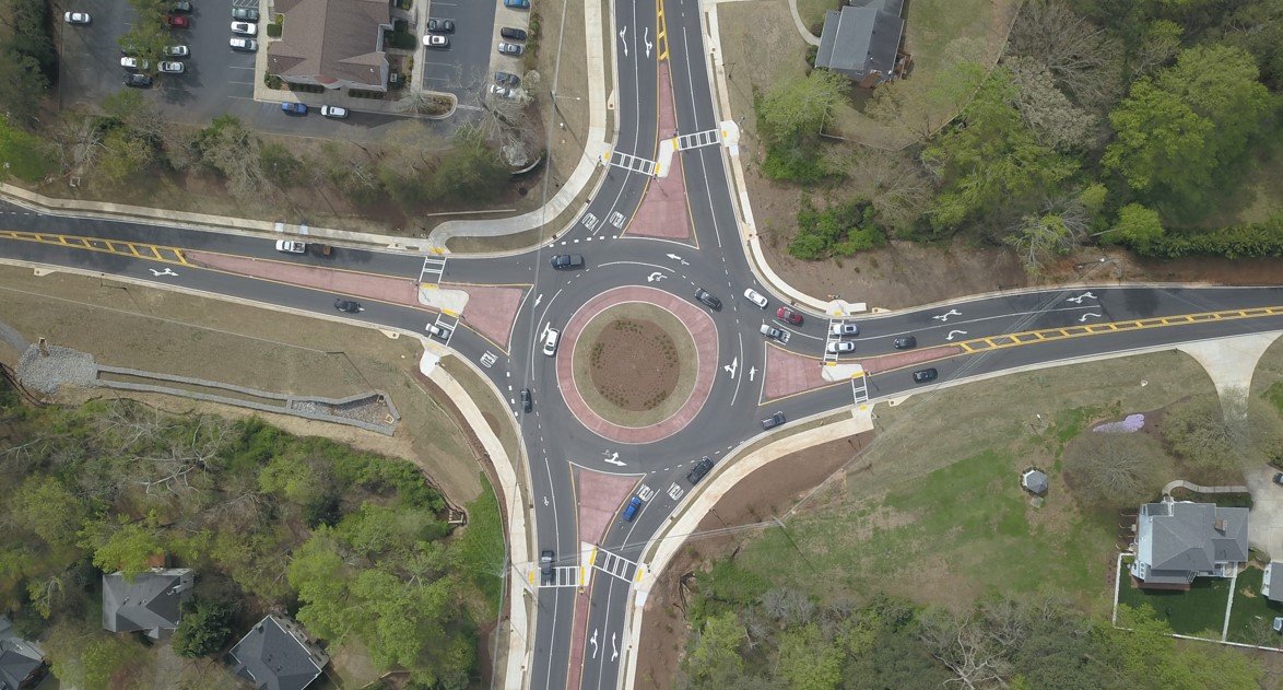 Aerial view of a four-leg, multi-lane modern roundabout. The design features a landscaped central island surrounded by a reddish mountable truck apron, raised splitter islands on each approach, high-visibility pedestrian crosswalks set back from the intersection, and pavement markings indicating yield lines and circulating lane directions.